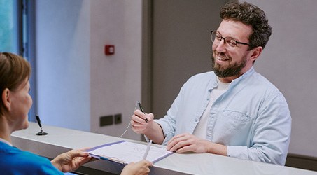 a dental patient filling out new patient forms being handed to him by a dental assistant
