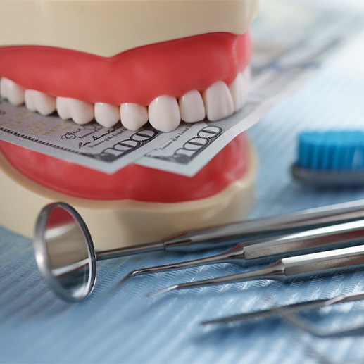 A jaw mockup biting some hundred-dollar bills next to some dental tools