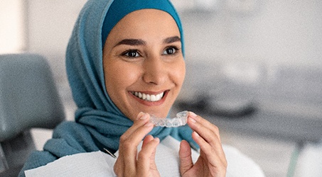 Woman smiling while holding Invisalign aligner in dental office